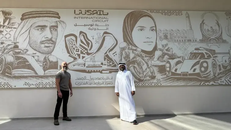 Artista vestido de negro e com barba comprida ao lado de um homem com uma jalaba vestida em frente a um painel em baixo-relevo com motivos alusivos ao mundo árabe e ao automobilismo